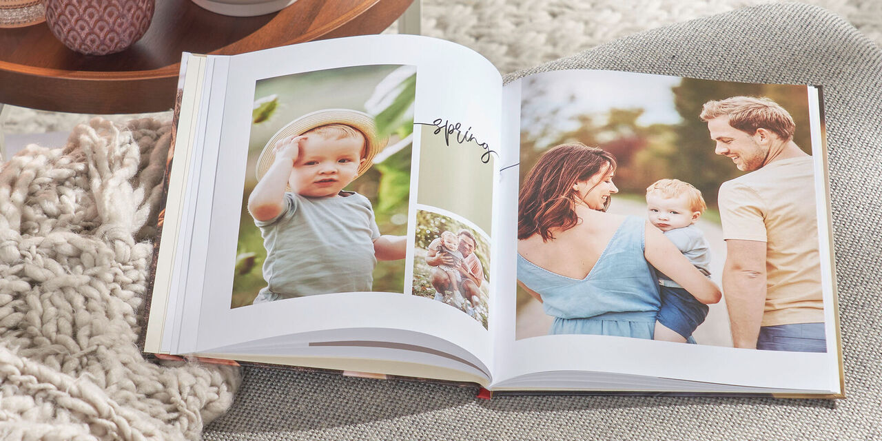 Auf einem grauen Sofa liegt ein aufgeschlagenes CEWE FOTOBUCH. Darin sind mehrere Fotos einer Familie sowie das Wort „Spring“ zu sehen. Links daneben steht ein Tisch mit Teelichtern und einer Pflanze.