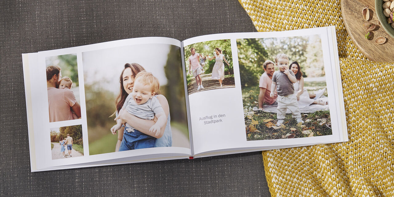 Auf einem Sofa liegt ein aufgeschlagenes CEWE FOTOBUCH, das mit dem Stil „Our Family“ gestaltet wurde. Auf der Doppelseite sind mehrere Motive einer Familie im Garten zu sehen. Rechts neben dem Fotobuch steht eine Schüssel Pistazien.