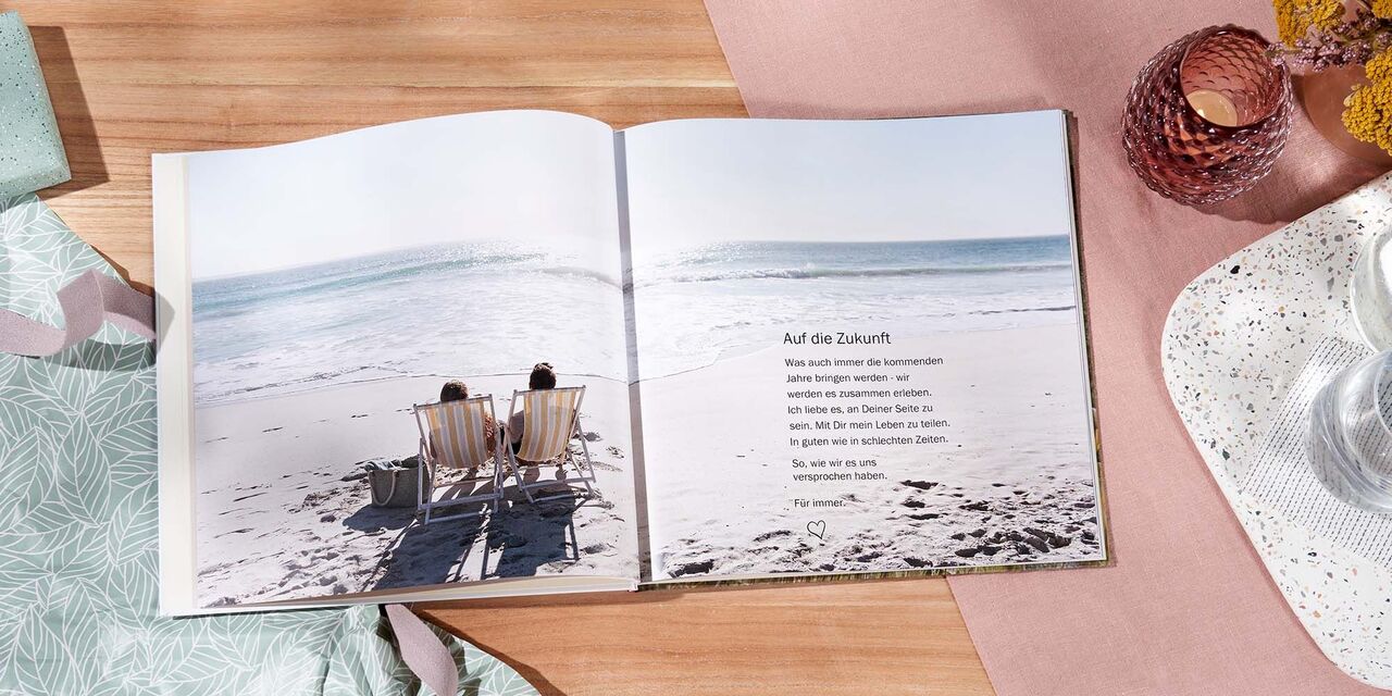 Auf einem Tisch liegt ein aufgeschlagenes CEWE FOTOBUCH. Über beide Seiten erstreckt sich die Aufnahme von einem Paar am Meer. Die Überschrift lautet „Auf die Zukunft“.