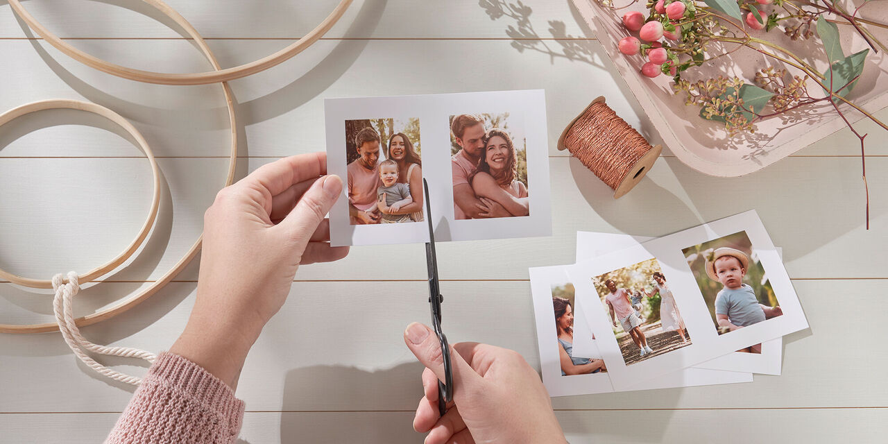 Aufnahme von zwei Händen, die ein CEWE Sofortfotoset mit zwei Bildern zuschneiden. Im Hintergrund liegen die weiteren Utensilien zum Basteln des Flower Hoops.