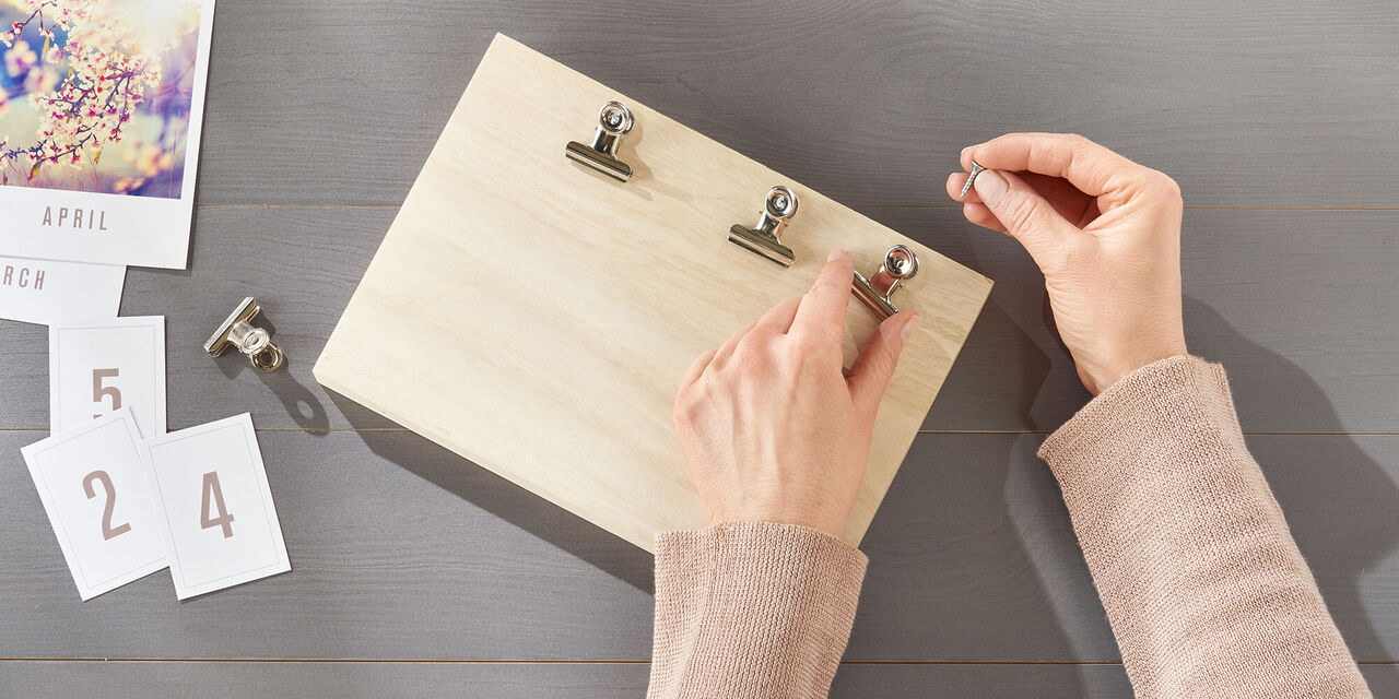 Ein Holzbrett liegt auf einem Tisch. Zwei Hände bringen drei Klemmen im oberen Bereich des Bretts an. Links liegen die ausgedruckten Fotos.