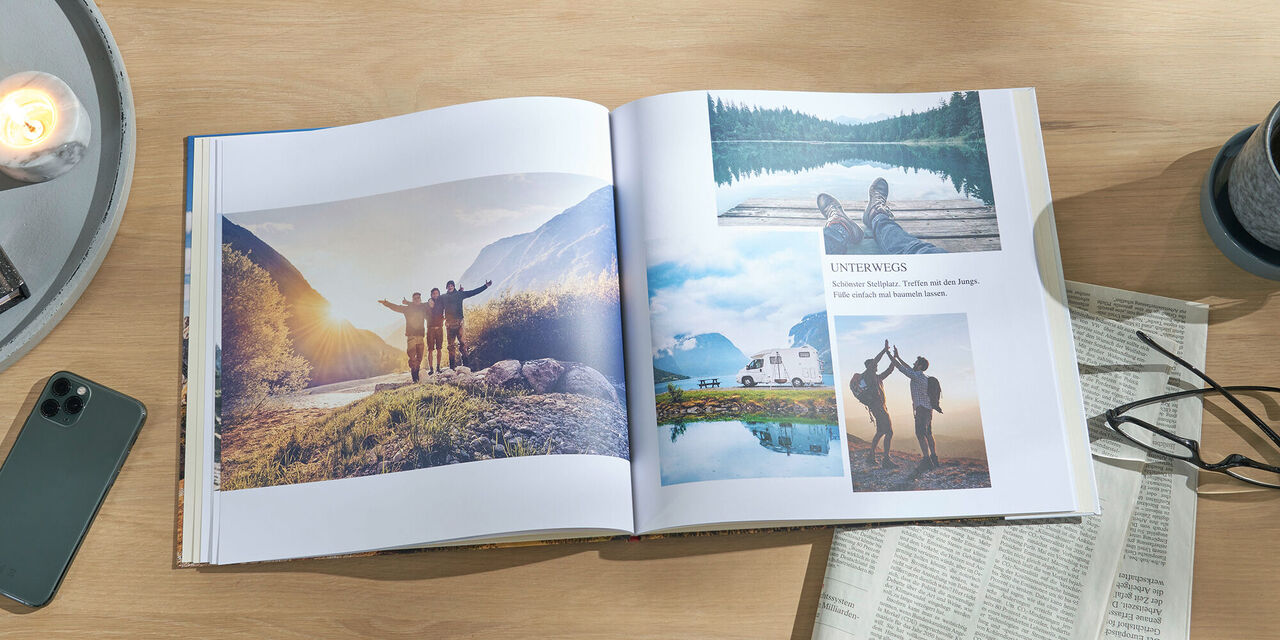 Ein aufgeschlagenes CEWE FOTOBUCH liegt auf einem Tisch. Darin sind mehrere Aufnahmen von Wanderern und der Natur zu sehen. Rechts daneben steht ein Becher mit Tee und es liegen eine Zeitung und eine Brille auf dem Tisch.