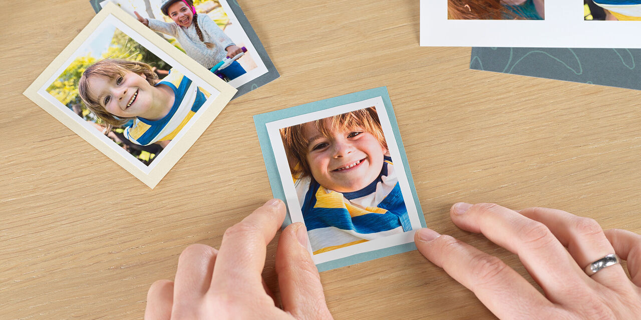 Nahaufnahme von zwei Händen, die ein Sofortfoto auf ein Rechteck aus Pappkarton kleben. Im Hintergrund sind weitere Sofortfotos zu sehen.