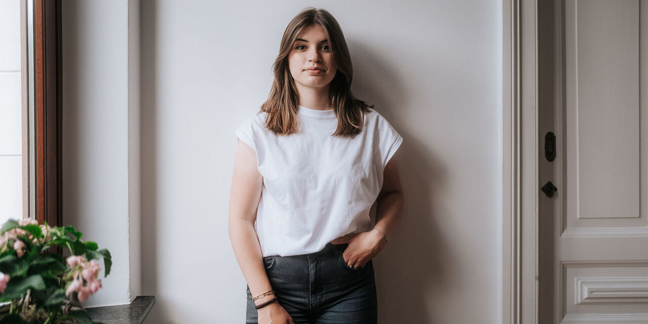 Eine Frau steht an einer Wand angelehnt und schaut in die Kamera. Sie hat dunkle Haare, trägt ein weißes T-Shirt und eine graue Jeans und hat eine Hand in ihrer linken Hosentasche.