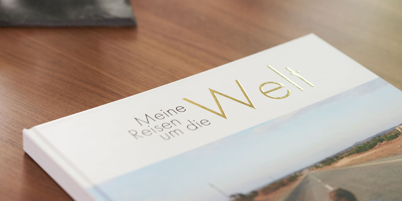 Auf einem Holztisch liegt ein CEWE FOTOBUCH, welches man nur im Anschnitt erkennt. Darauf zu sehen ist Lea Wippermann in der Mitte einer großen leeren Straße. Das "Welt" im Titel des Fotobuchs ist mit Gold veredelt. Oben links im Bild sieht man die Ecke eines schwarzen Tabletts.