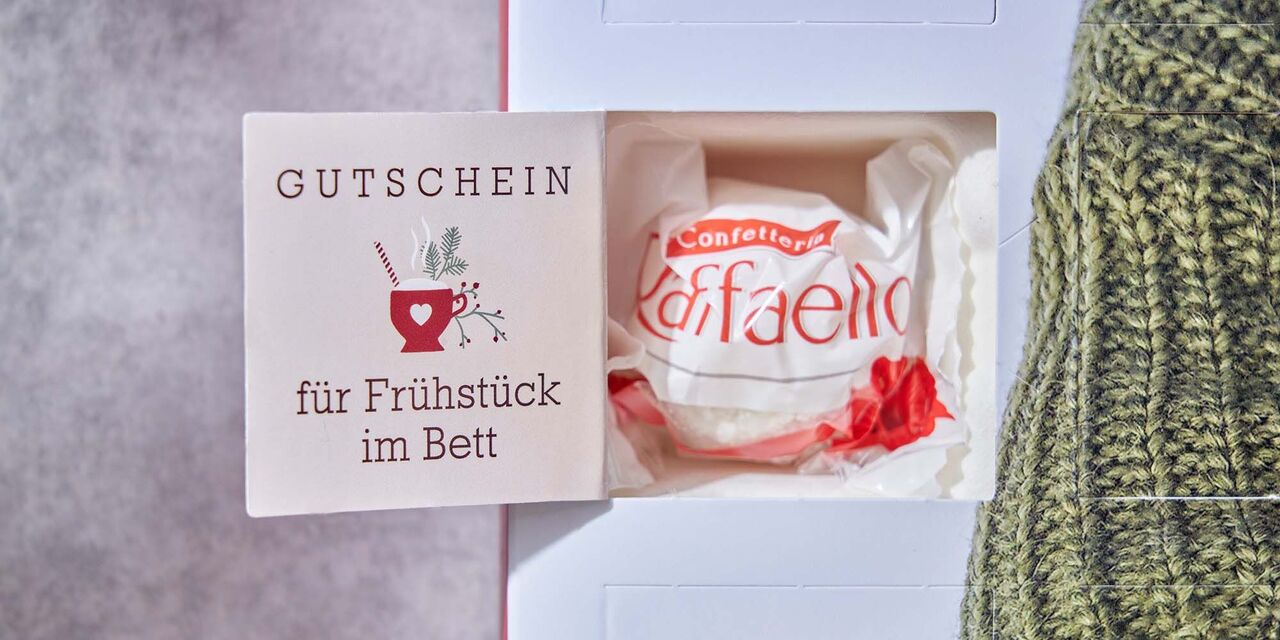 Auf der Innenseite eines geöffneten Adventskalendertürchens steht der Text “Gutschein für Frühstück im Bett”. Außerdem befindet sich eine Tasse mit weihnachtlicher Deko als Designelement auf der Tür, hinter der eine Raffaello-Praline steckt.