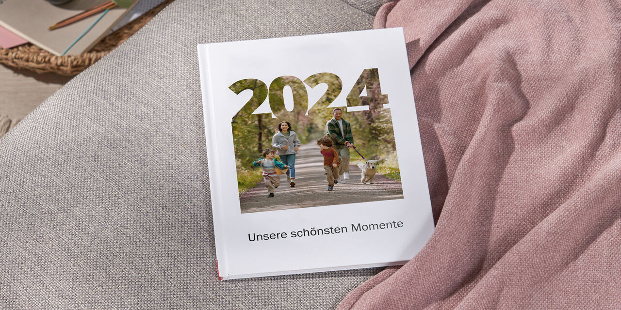 Auf einem Sofa liegt ein CEWE FOTOBUCH, das mit dem Stil „Best of the Year“ gestaltet wurde. Auf dem Titel sind ein Motiv einer Familie im Schnee sowie die Jahreszahl 2022 zu sehen. Rechts neben dem Fotobuch liegt eine rosafarbene Decke.