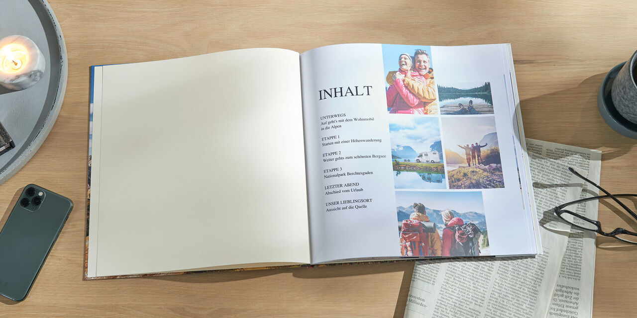 Ein aufgeschlagenes CEWE FOTOBUCH liegt auf einem Tisch. Darin sind Aufnahmen von Wanderern und ein Inhaltsverzeichnis zu sehen. Rechts daneben steht ein Becher mit Tee und es liegen eine Zeitung und eine Brille auf dem Tisch.